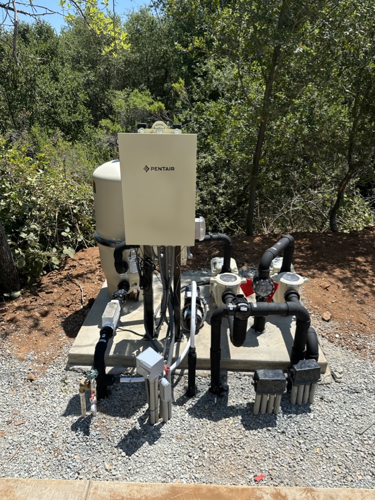 Outdoor pool equipment: a Pentair control cabinet with pumps, filters, and insulated pipes on a concrete pad in a wooded area.