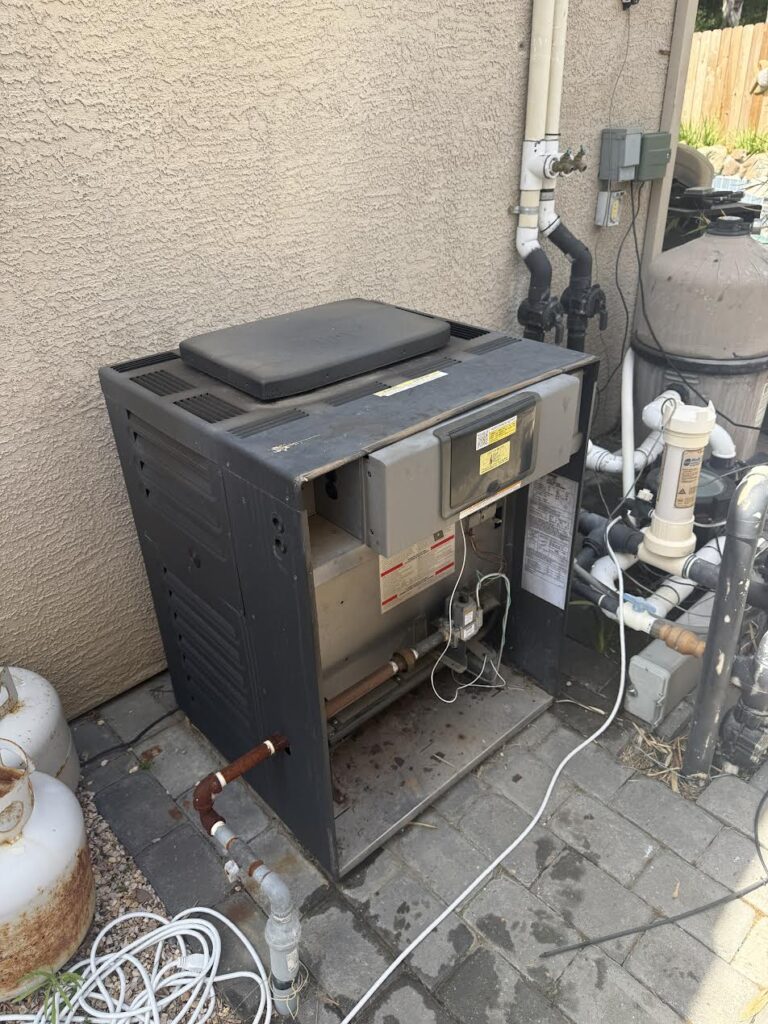 Outdoor heating/AC unit with open access panel and exposed wiring beside a stucco wall, surrounded by pipes and a propane tank outside.