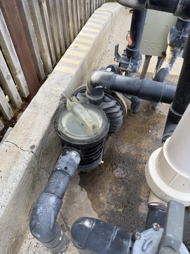 Outdoor pool filtration pump with PVC piping and valves, set on a damp concrete surface beside a wooden fence.