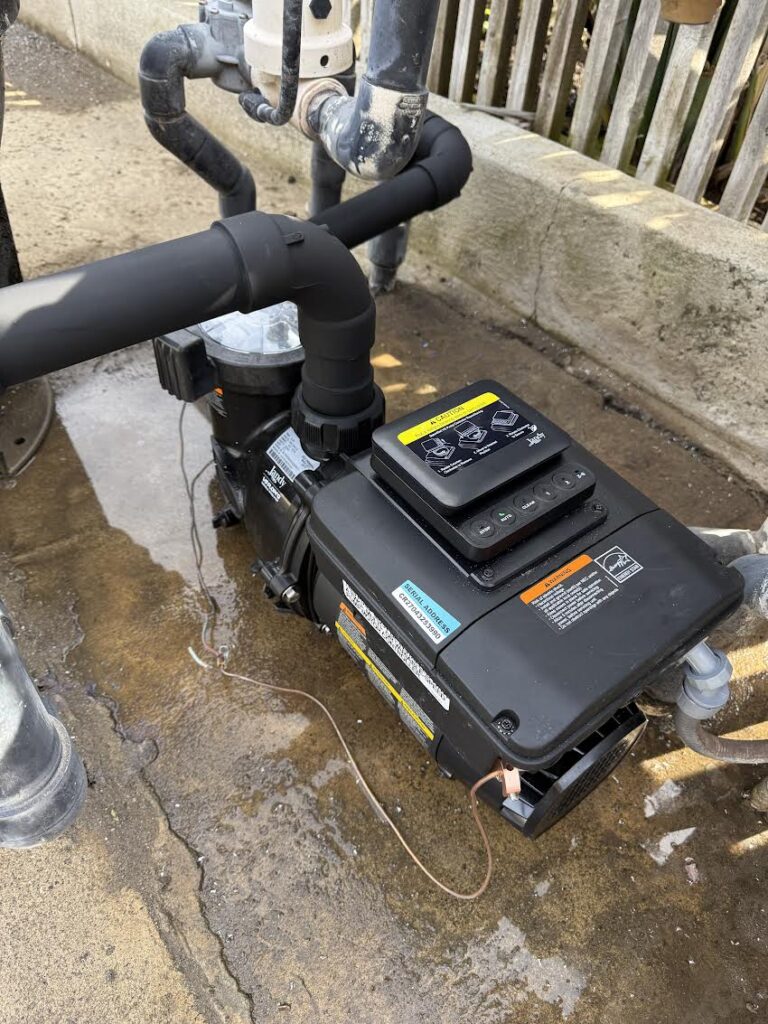 Above-ground pool pump with black insulated pipes and electrical control box, sitting on wet concrete near a fence.