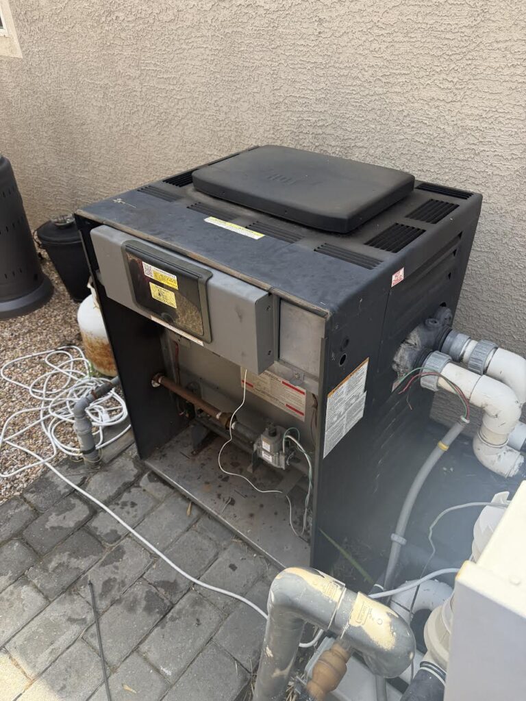 Outdoor HVAC unit (furnace/air conditioner) with exposed wiring and copper pipes beside a stucco wall on a paved area.