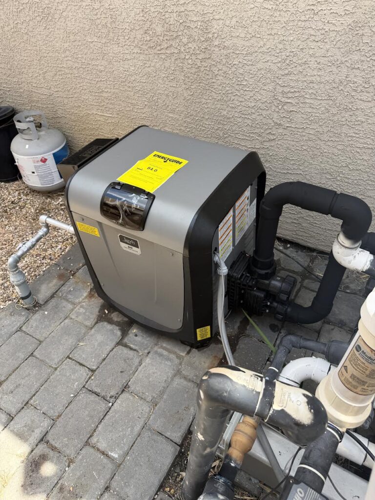 Outdoor pool heat pump with a yellow energy guide label, connected to PVC piping along a textured exterior wall and adjacent propane tank.