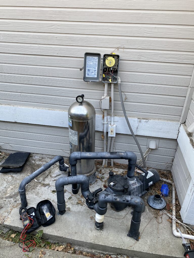 Outdoor pool equipment with a pump, pipes, and a gauge mounted on a wall behind a concrete slab.