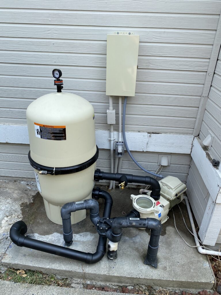 Outdoor pool filtration system with a tall beige filter tank, pressure gauge on top, and a network of black PVC pipes along a siding wall.