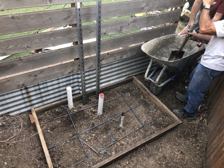 Foundation trench at a construction site with rebar grid, PVC pipes, and a worker shoveling concrete into a wheelbarrow