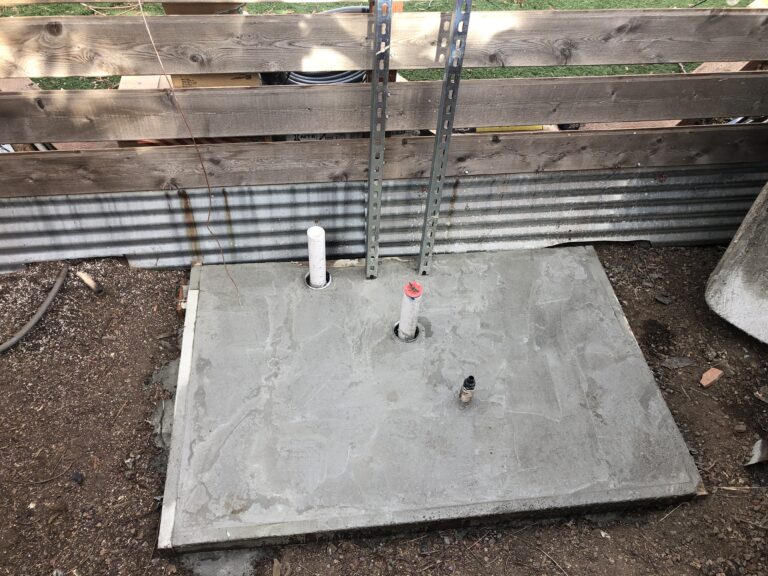 Concrete slab with two vertical plumbing pipes and metal framing studs at a construction site set behind a wooden barrier.