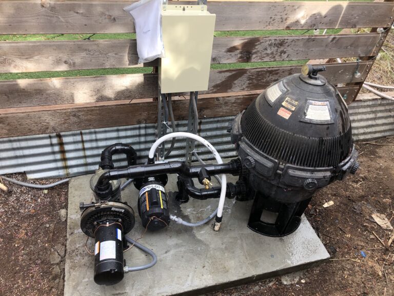 Outdoor pool pump and filter system with PVC piping on a concrete pad beside a weathered wooden fence.