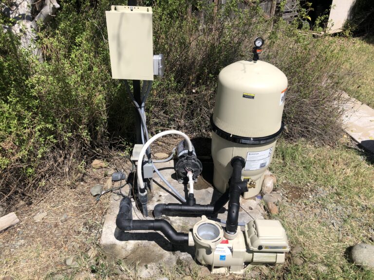 Outdoor pool filtration system with a tan cylindrical tank, pressure gauge on top, and connected pipes and electrical box nearby.
