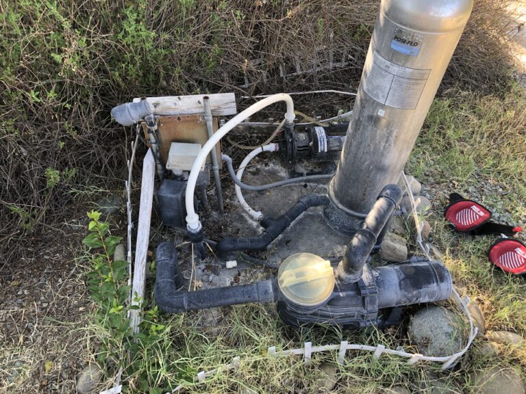 Outdoor pump system with hoses, pipes, and a large vertical tank surrounded by dry, overgrown brush.