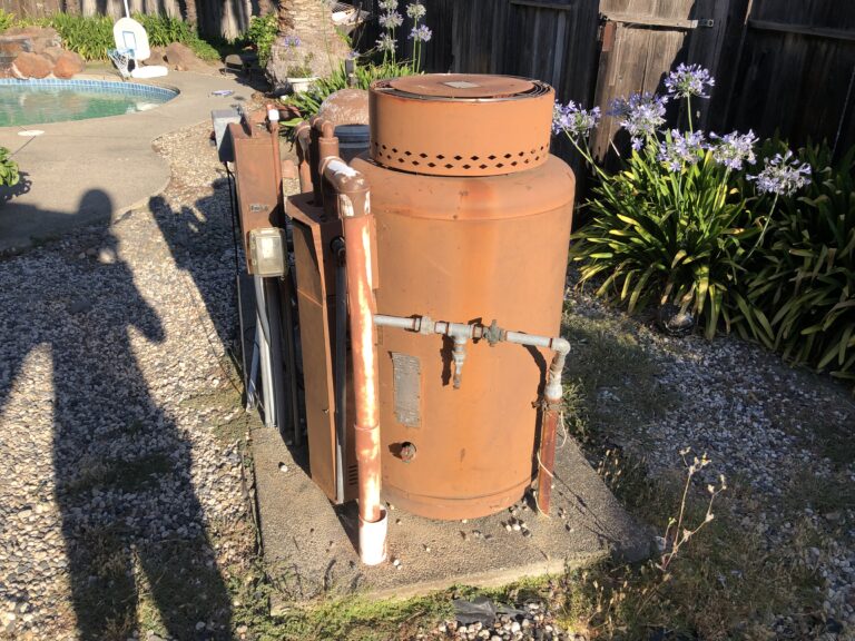 Large rusty orange propane tank with connected pipes in a gravel backyard beside a wooden fence and purple flowering plants by the garden bed, near a pool area distant in the background.
