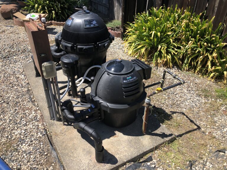 Outdoor backyard scene with a black round pool filter and pump system on a gravel surface beside green shrubs by a wooden fence.