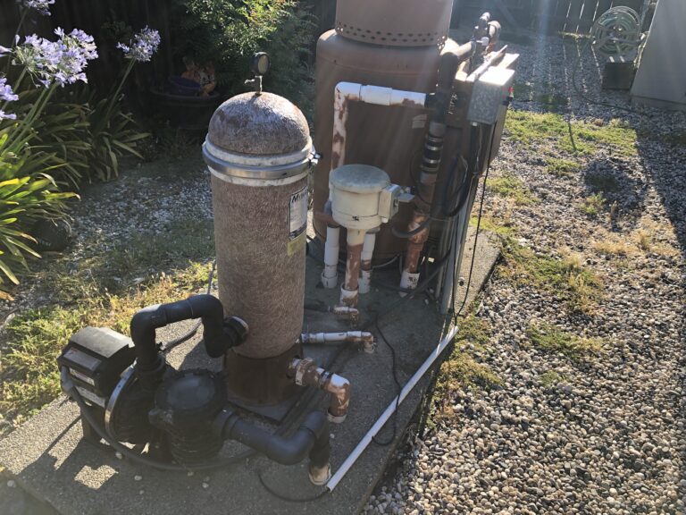 Outdoor pool heater/boiler apparatus with rusted metal tank, white PVC pipes, and black hoses in a gravel yard.