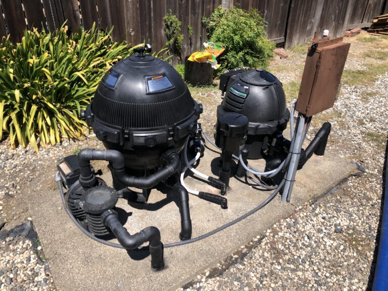Two large black swimming pool filters with attached hoses set up in a gravel backyard against a wooden fence, with plants nearby.