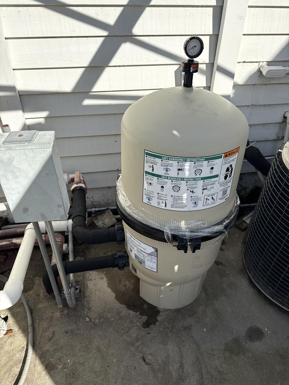 Above-ground pool filter unit with a pressure gauge on top, connected to hoses beside the house siding.