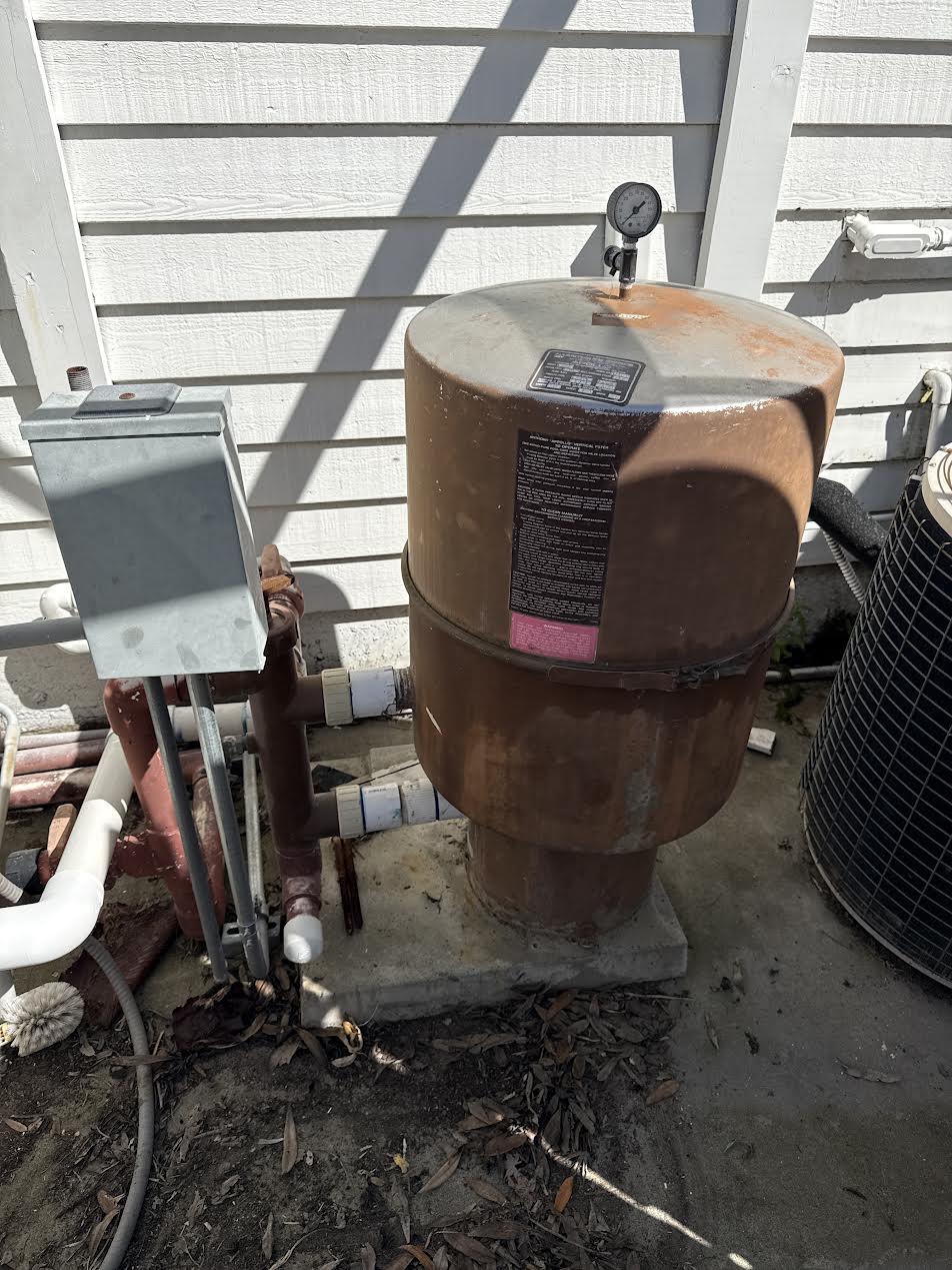 Outdoor rusty water pressure/expansion tank with top gauge beside a house, connected to plumbing pipes and an electrical box